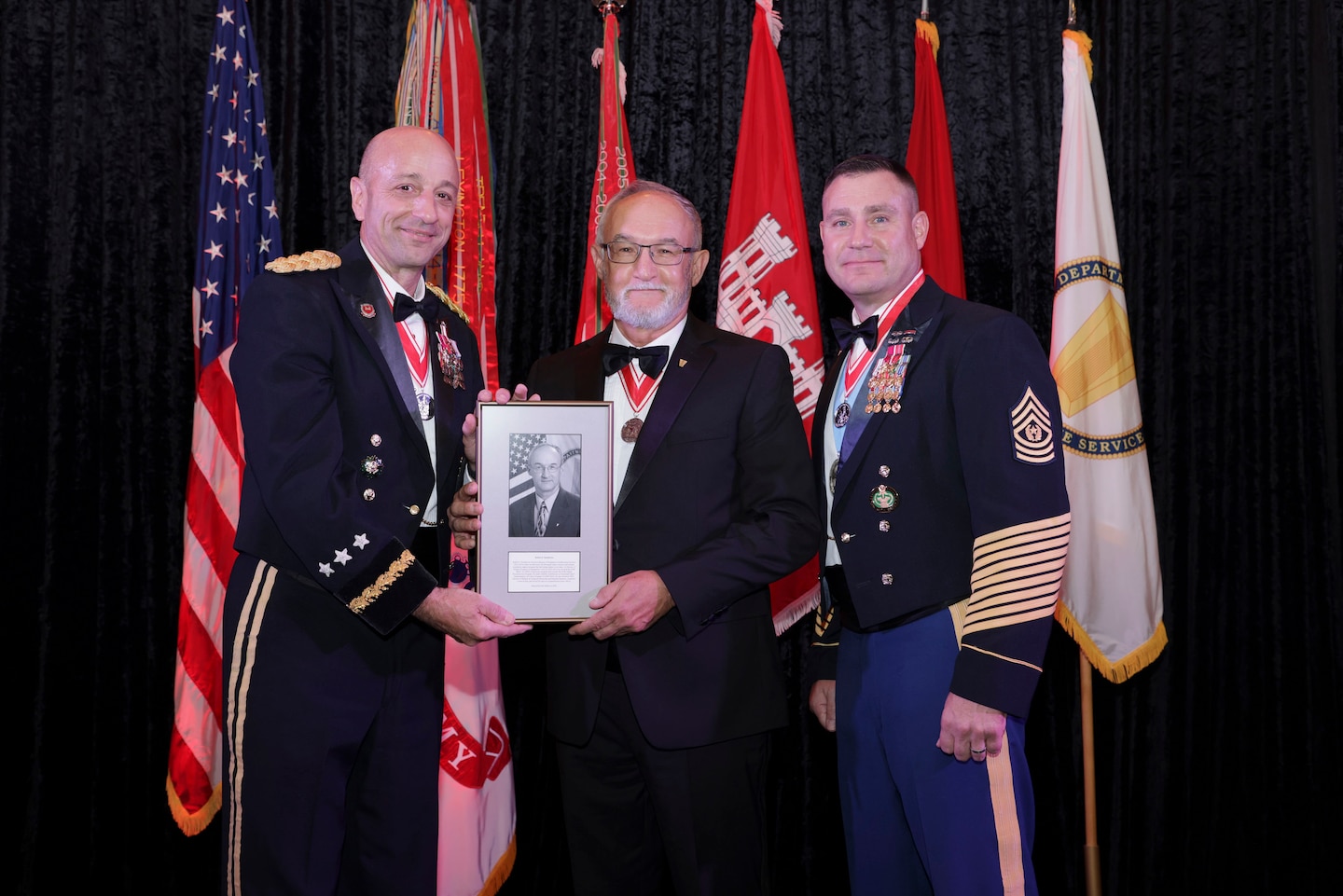 Three men stand in front of flags, the two men on each side are wearing Army dress uniforms, the man in the center is wearing a suite.