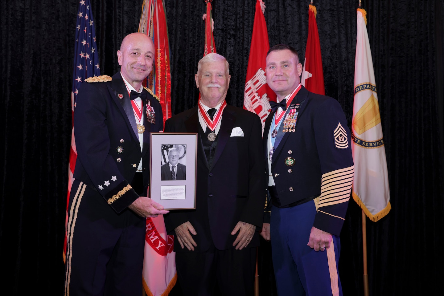 Three men stand in front of flags, the two men on each side are wearing Army dress uniforms, the man in the center is wearing a suite.