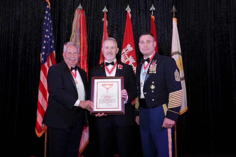 Three men stand in front of a row of flags. The gentleman on the right is in Army dress uniform the other two men are in tuxedoes. The man in the center is holding a plaque.