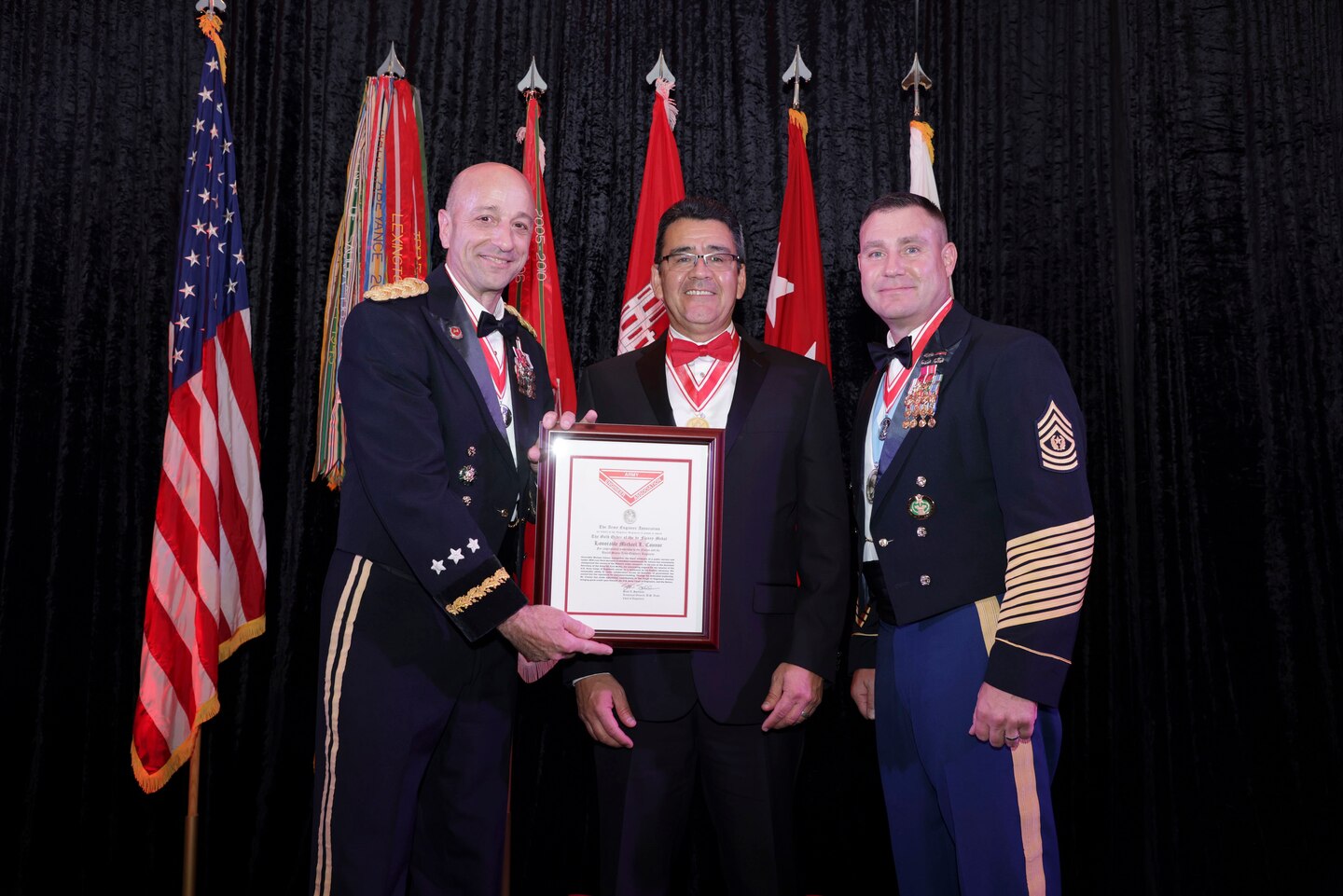 Two men in in dress uniform are on either side of a man in a tux as he is being presented a plaque.