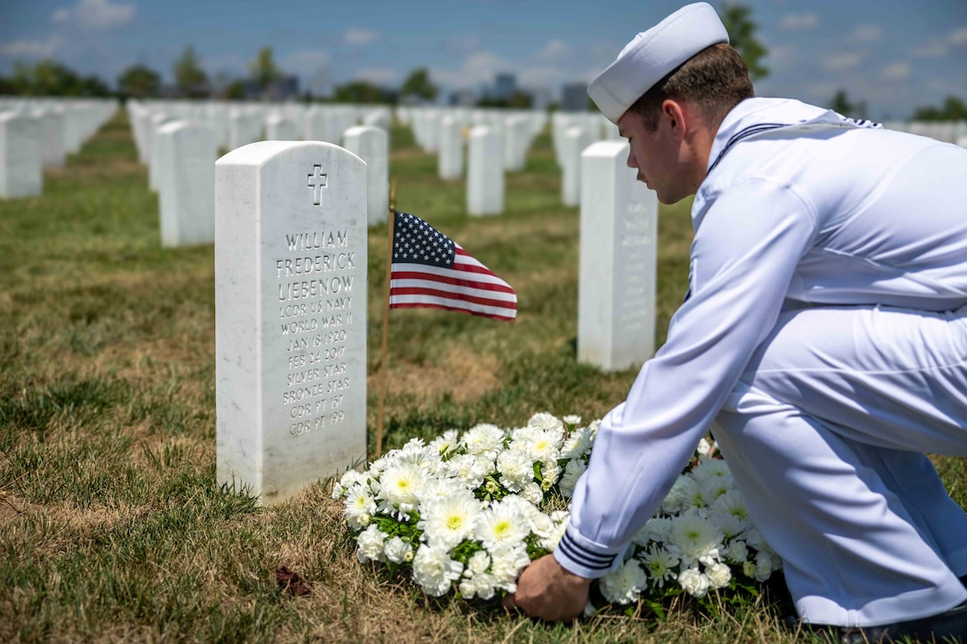 A sailor places a wreath of white flowers at the base of a gravestone with a small American flag next to it.