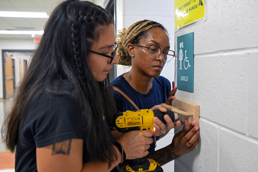 Two sailors use a cordless screwdriver to attach a small wooden coat hanger to a wall in a school.