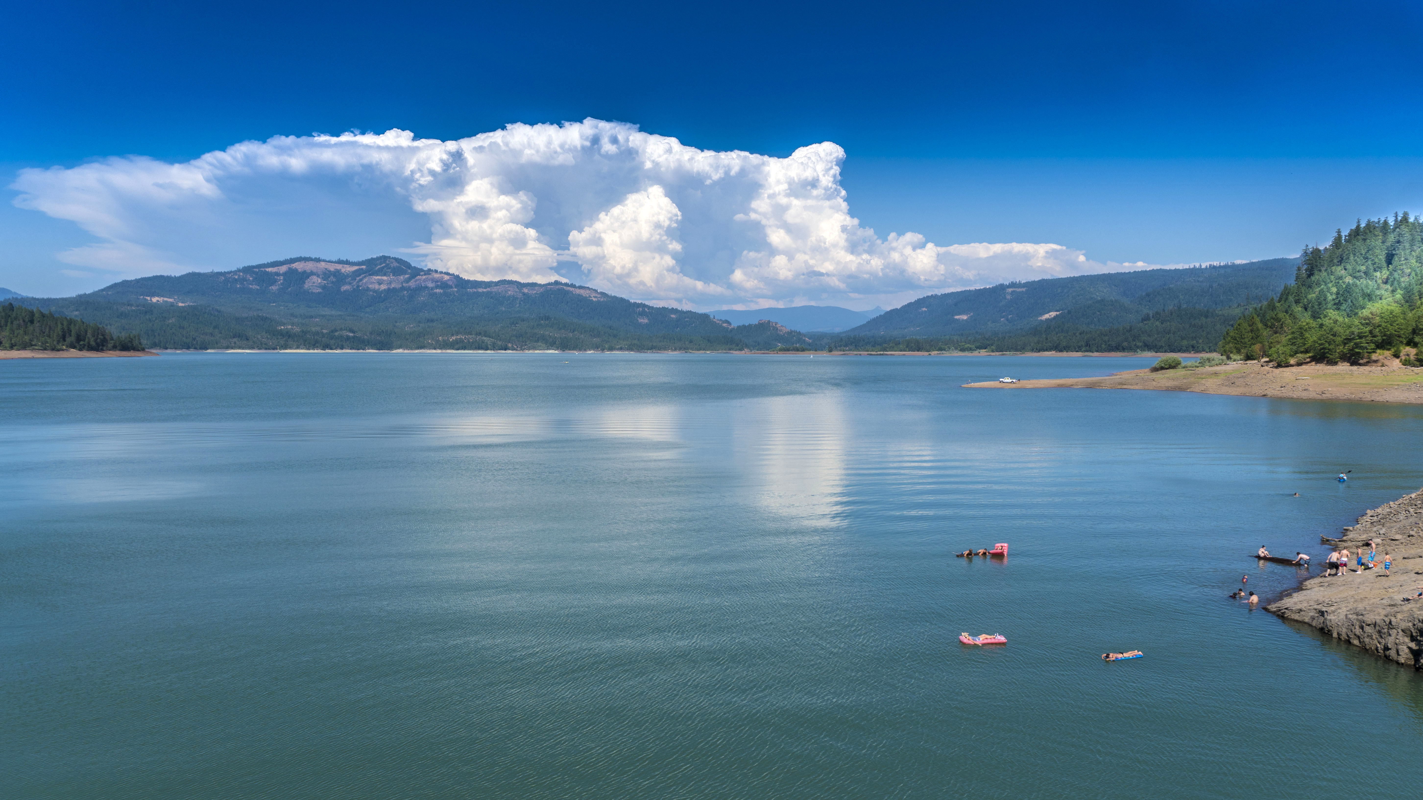 A group of people are on floats in the water at Lost Creek Reservoir. The shoreline, is visible. Beautiful mountains with clouds draped over them is in the background.