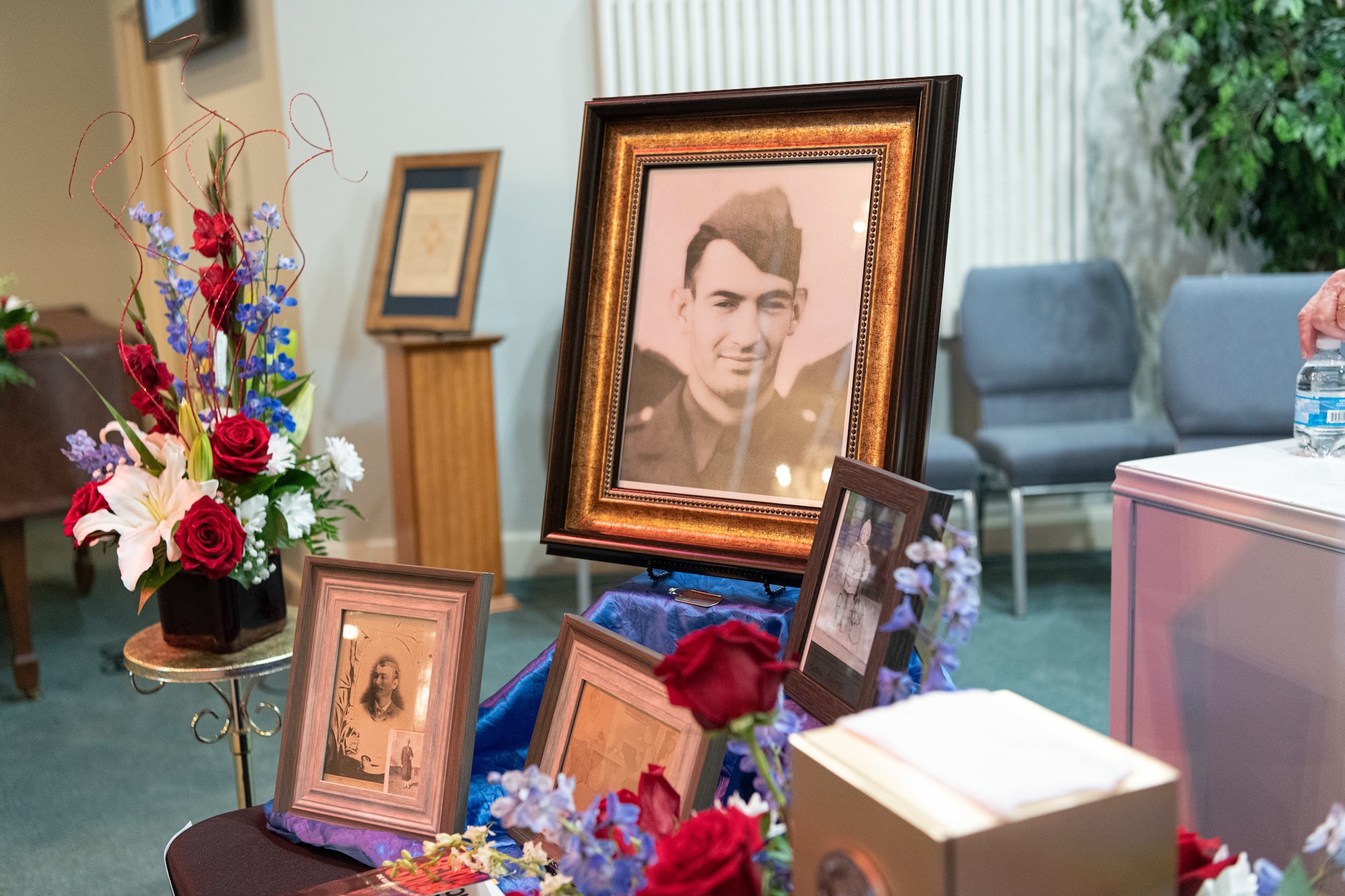 A black and white photograph of a man in military uniform stands on display next to several framed photographs and bouquets of flowers.