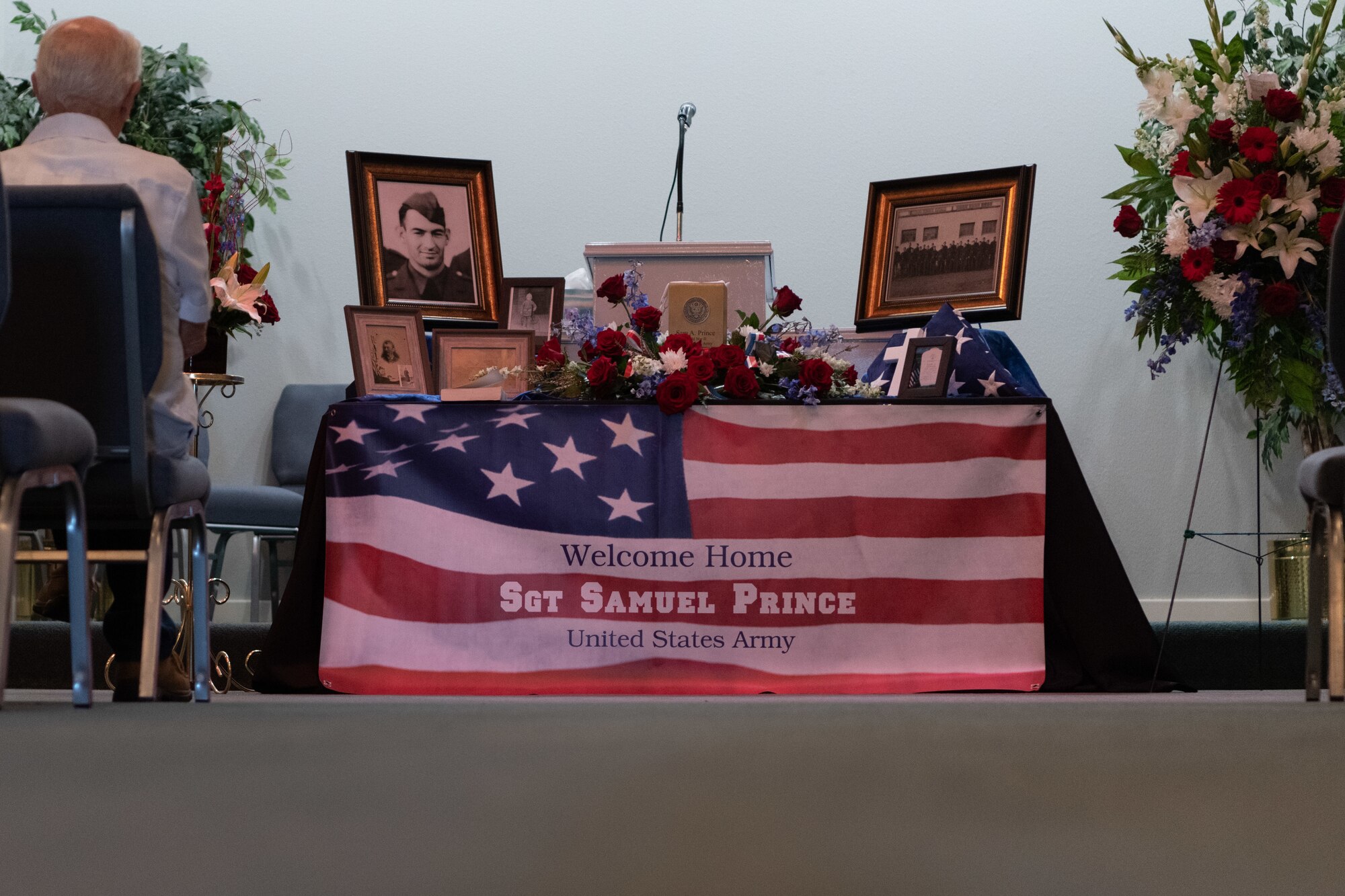Several people sit in front of a display table holding an American flag and several framed photographs.