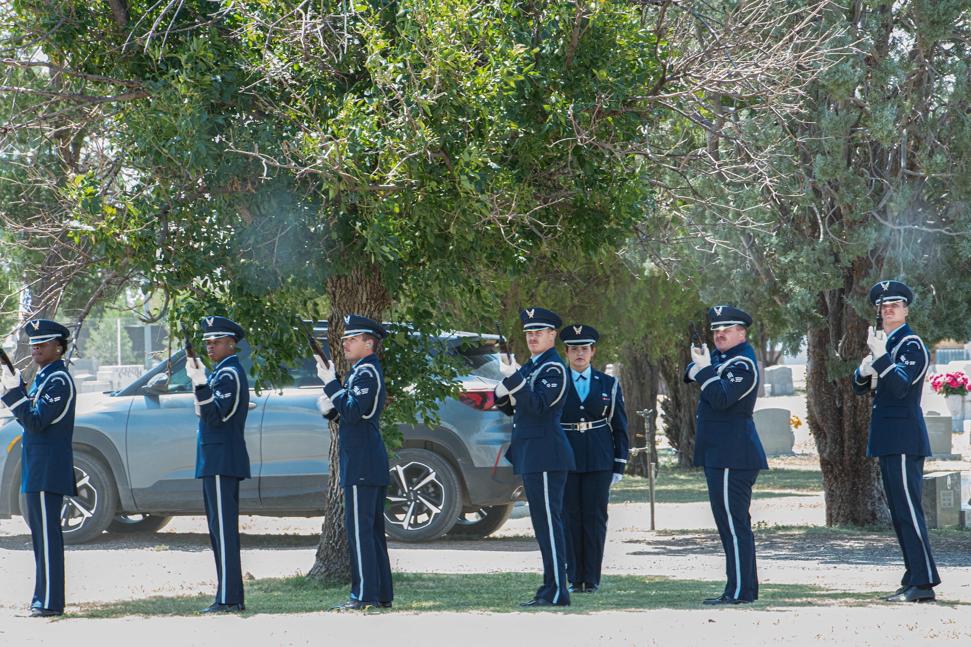 A line of men and women in military dress uniforms fire ceremonial rifles during a funeral event at an outdoor cemetery.