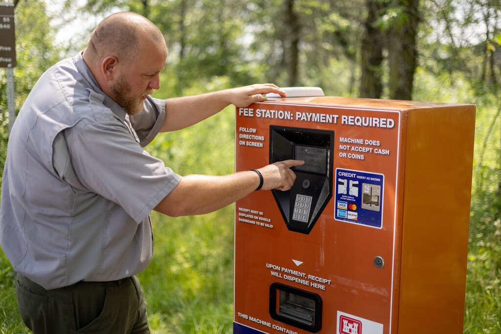 A man in a grey shirt and green pants touches the keypad of an orange machine with greenery in the background.