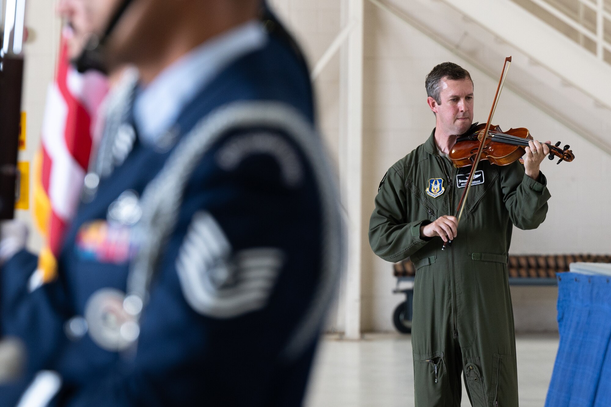 A man in a U.S. Air Force flight suit plays a violin. In the foreground, the blurred uniform of a U.S. Air Force Honor Guard member.