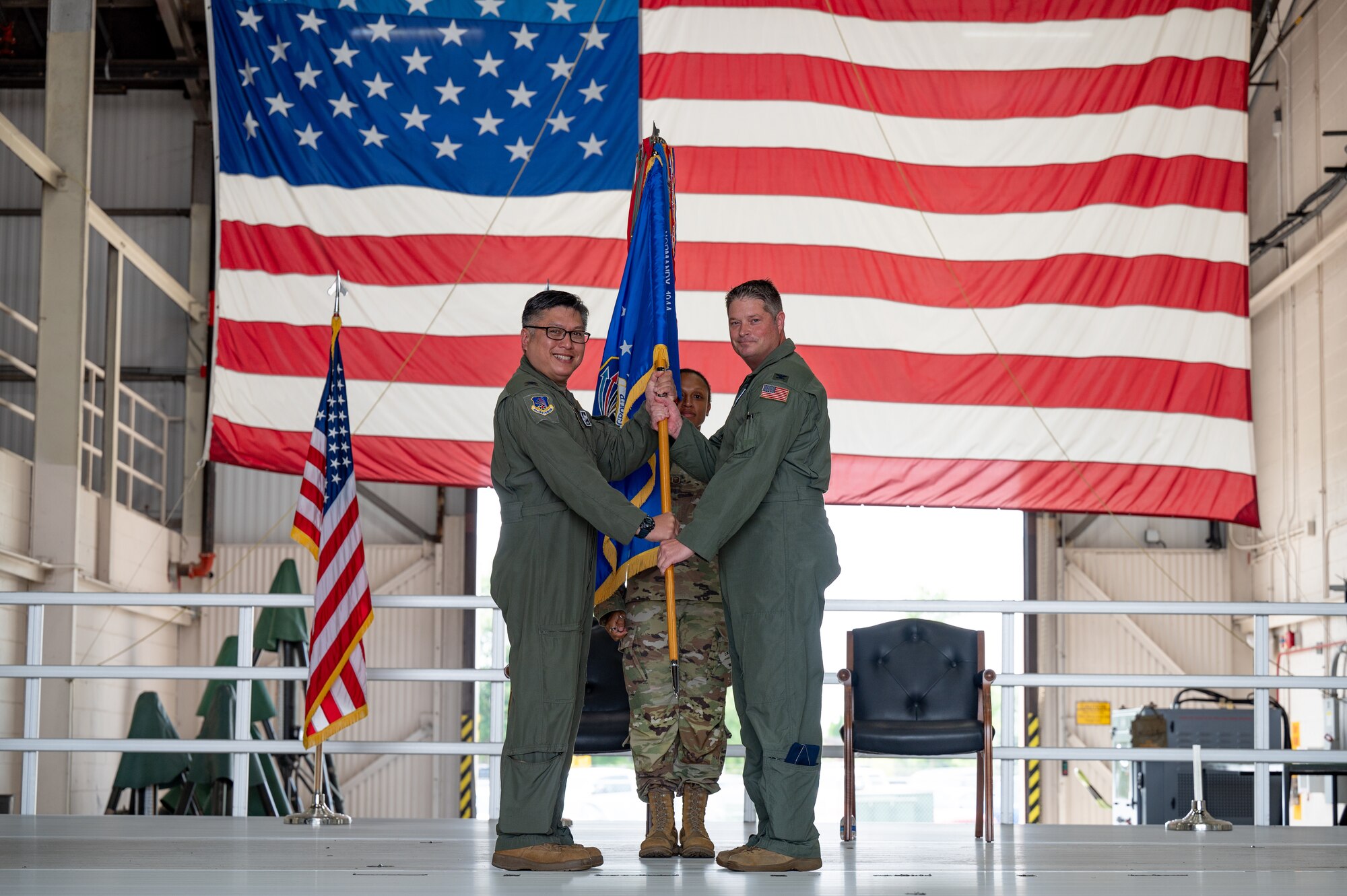 A man in a U.S. Air Force flight suit smiles while holding a guidon, passing the flag to another man in a U.S. Air Force flight suit. Both men are posed, holding onto the guidon, for the photo.