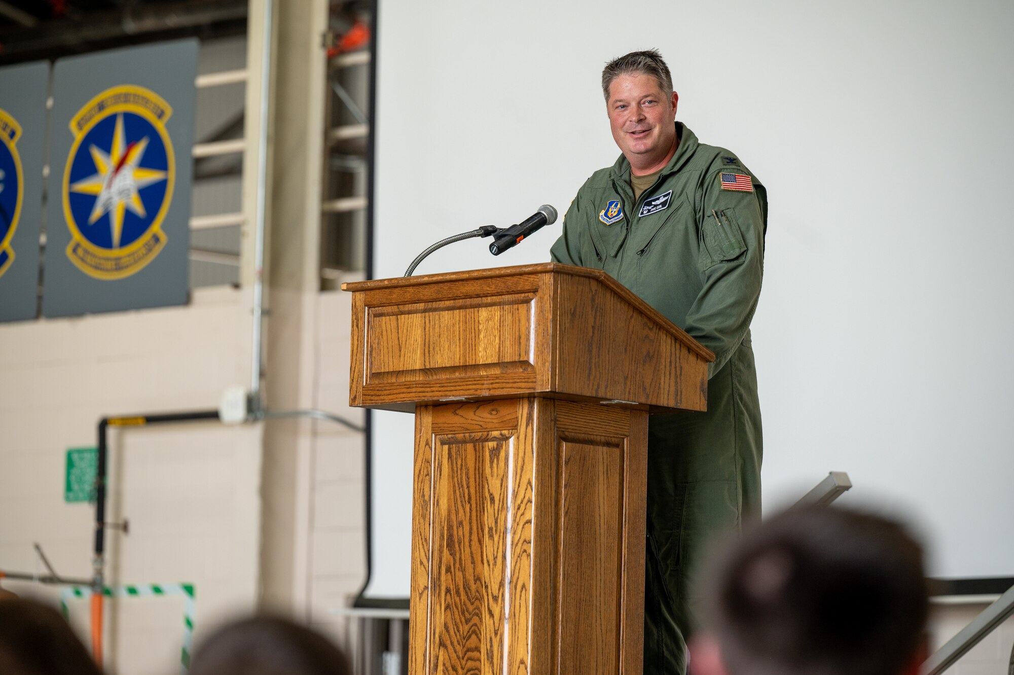 A man in a U.S. Air Force flight suit standing behind a wooden podium and smiling into a crowd. The tops of the audience's heads are visible yet blurred.