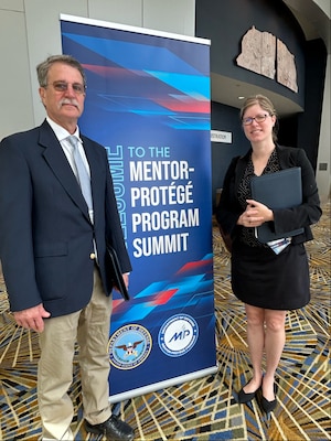 A man and woman stand on either side of a poster advertising DOD's Mentor-Protege Program Summit.