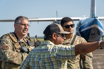 With a large aircraft in the background, two men in uniform speak to a man in civilian attire. The civilian man is pointing towards something in the distance and the two uniformed men are looking that direction.