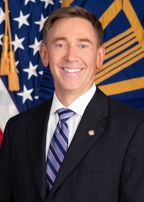 Kenneth Watson, Deputy Commander, Defense Logistics Agency Land and Maritime, man in a dark suit with flags behind him. He is smiling.