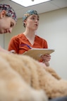 A veterinarian in a red uniform is holding papers while talking to other people nearby. There is a large, light brown dog in the foreground laying on a table.