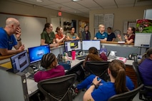 Doctors and nurses in various colors of hospital uniforms are standing and sitting around a nurses' station in an animal hospital discussing something.