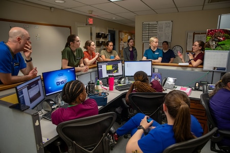 Doctors and nurses in various colors of hospital uniforms are standing and sitting around a nurses' station in an animal hospital discussing something.