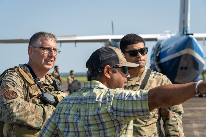 With a large aircraft in the background, two men in uniform speak to a man in civilian attire. The civilian man is pointing towards something in the distance and the two uniformed men are looking that direction.