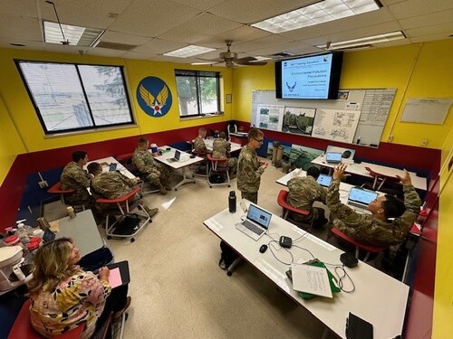 A woman watches a group of Airmen in a classroom.