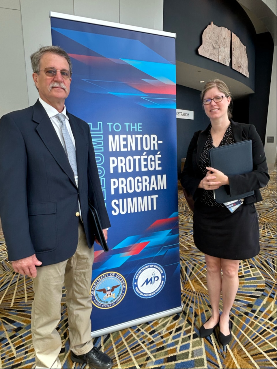 A man and woman stand on either side of a poster advertising DOD's Mentor-Protege Program Summit.