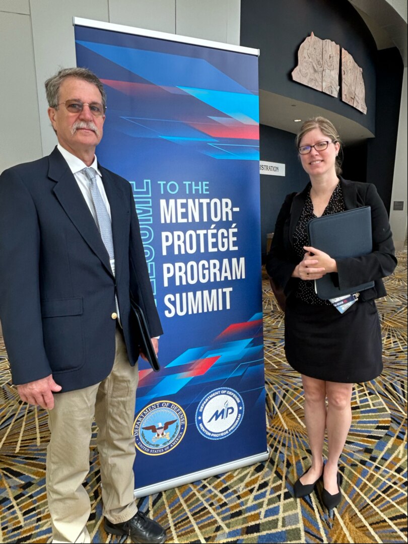 A man and woman stand on either side of a poster advertising DOD's Mentor-Protege Program Summit.
