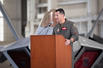 Lt. Col. Peter “Sly” Liggieri, incoming 149th Fighter Squadron commander, stands at a podium with an F-22 Raptor in the background.