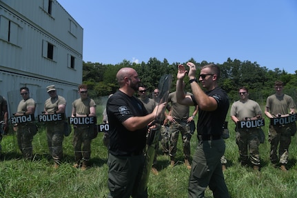 Two police officers demonstrating techniques to line of Airmen holding police shields behind them.