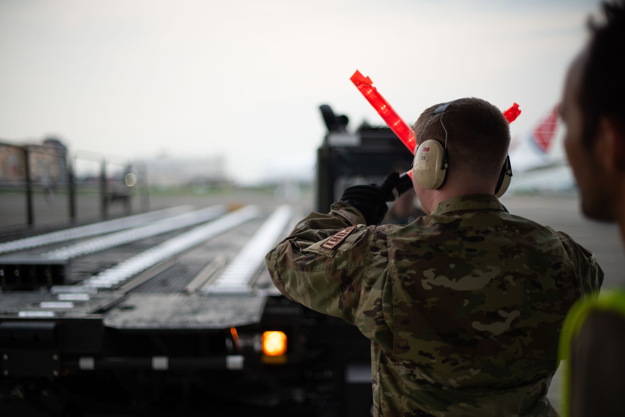A U.S. Air Force Airman uses red marshalling wands to guide a large platform vehicle.