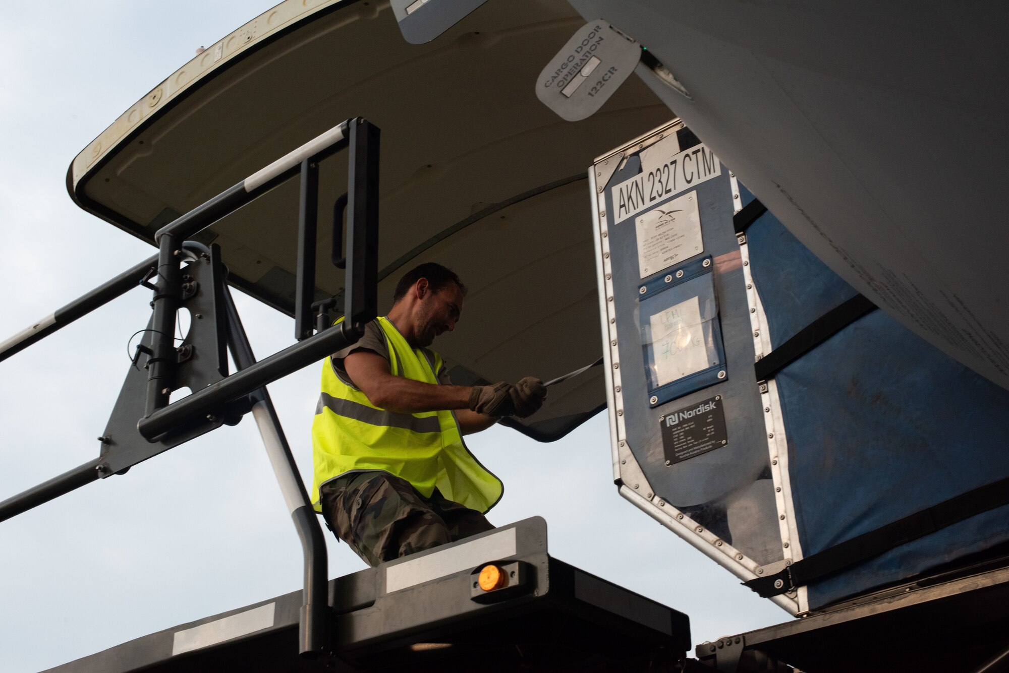 A man in a high-visibility vest pulls a large crate out of an aircraft onto a platform.