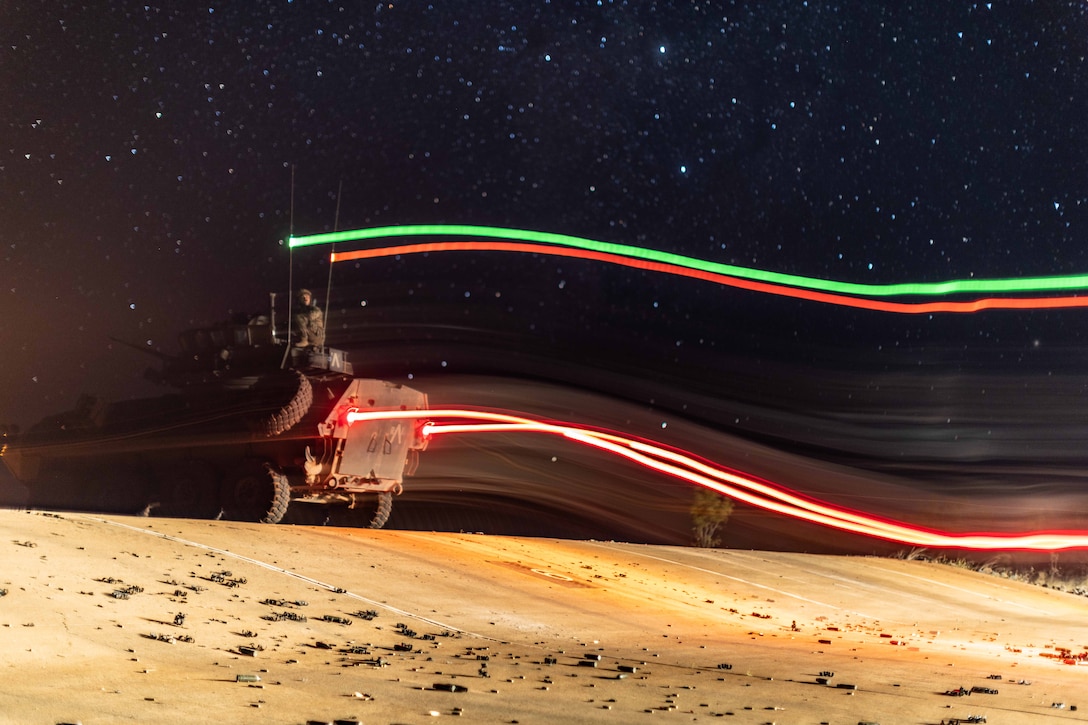 Streaks of multicolored lights emanate from different parts of a military vehicle as it sits in a desert area at night.