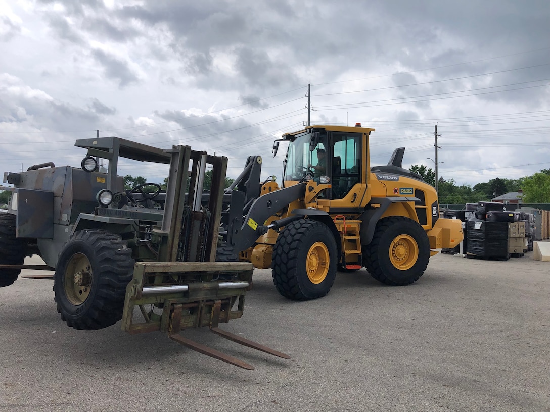 A large yellow piece of equipment has lifted an olive green forklift and is moving it across a parking lot.