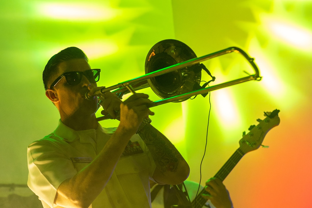 Close-up of a sailor playing a trombone during a performance with yellow, green and orange lights in the background.