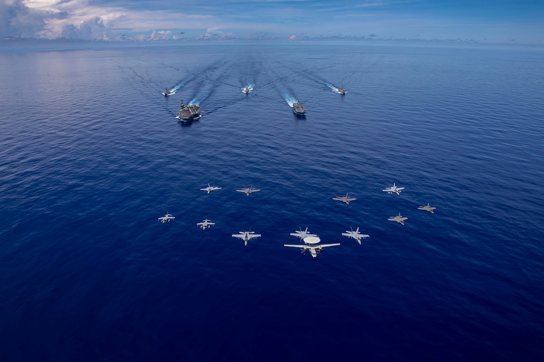 Five U.S. and foreign ships sail in open water during daylight as a dozen military aircraft fly ahead.