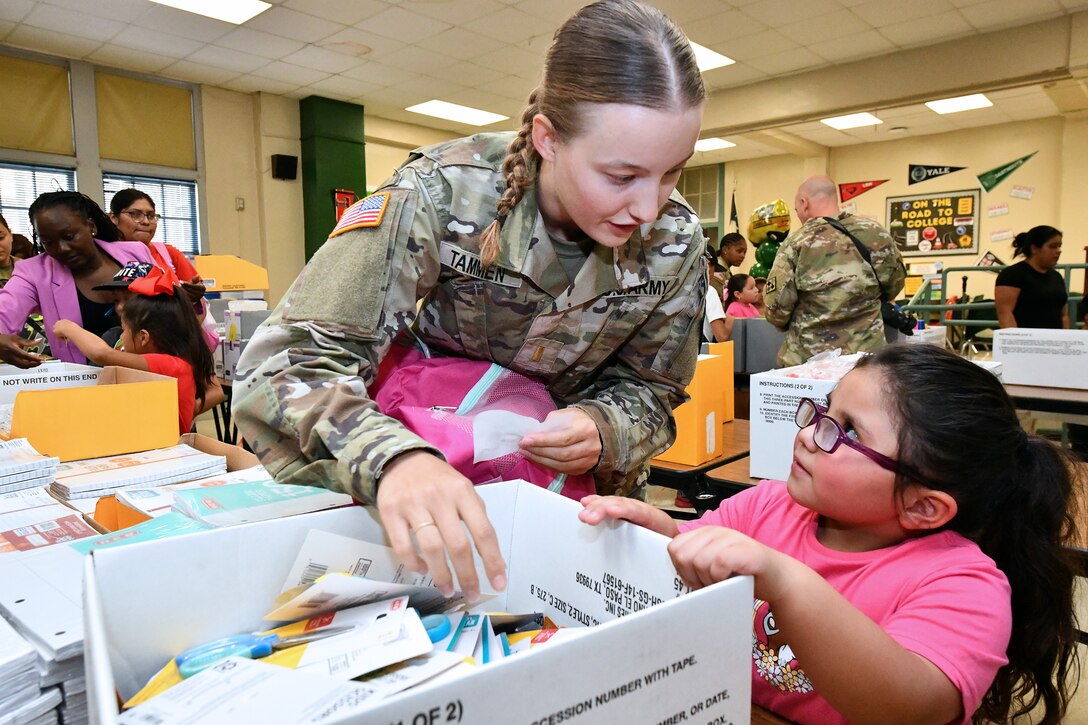 A soldier leans to talk to a child while distributing scissors from a box during a back-to-school supply drive with other service members, civilians and students in the background.