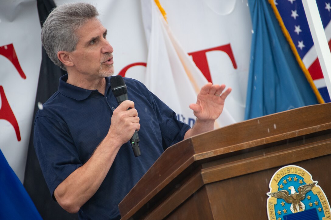 A man in a blue polo shirt speaks into a microphone behind a podium.