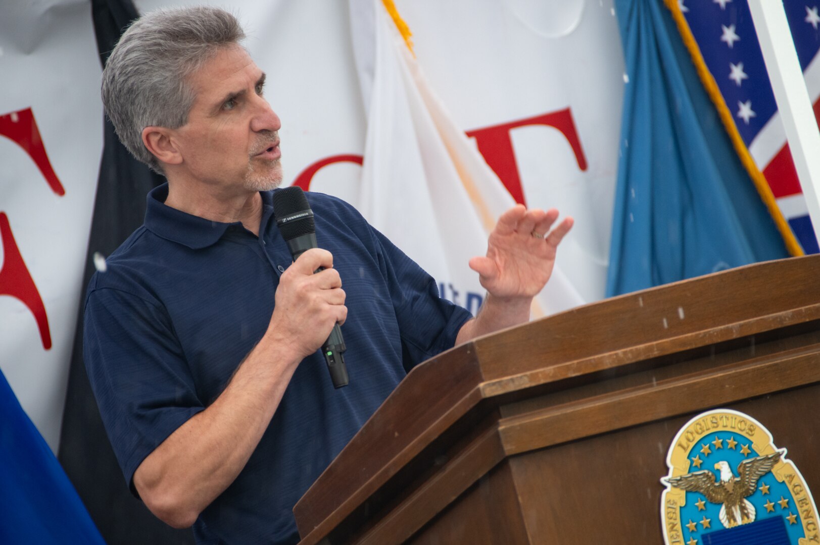 A man in a blue polo shirt speaks into a microphone behind a podium.