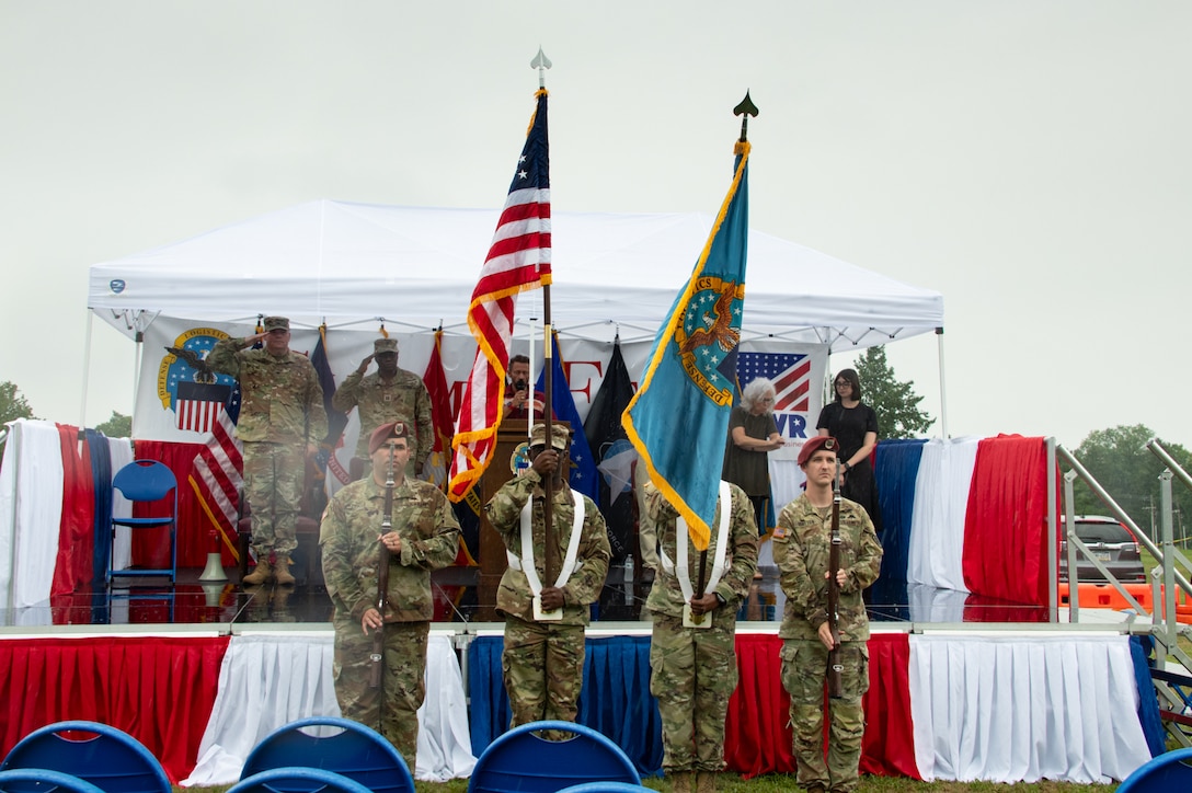 Four Army soldiers dressed in camouflage stand in front of a stage. The two in the middle are holding an American flag and the Defense Logistics Agency flag.