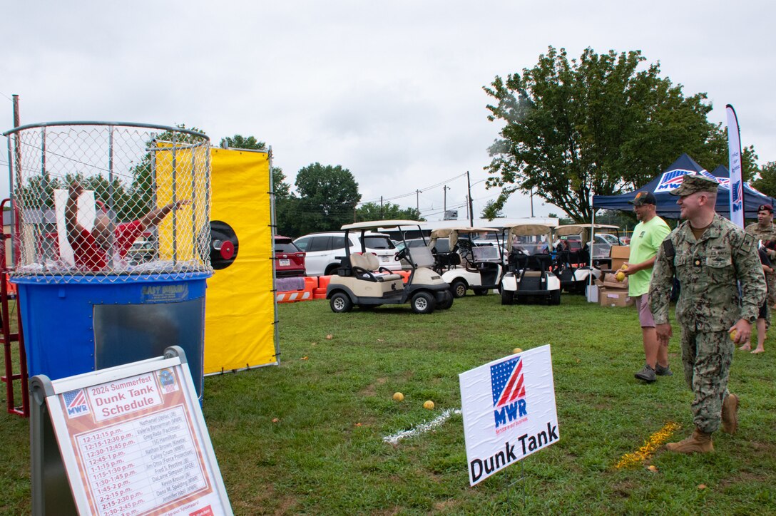 A Navy officer in camouflage throws a ball at a dunk tank, dunking the man in the tank
