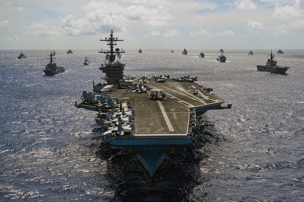 Viewed from above, an aircraft carrier sails ahead of a group of military ships at sea during a sunny day.