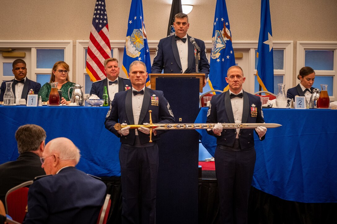 Chief Master Sgt. Gregg Gow, serving as Chief Master Sergeant of the Mess, addresses the audience from the podium during the OSI Order of the Sword ceremony, July 31, 2024, at Marine Corps Base Quantico, Virginia. From right: Chief Master Sgts Charles Pipkins and James Hoy, serving as Keeper of the Sword and Guardian of the Sword respectively, carefully place the hand-painted OSI sword in its place.