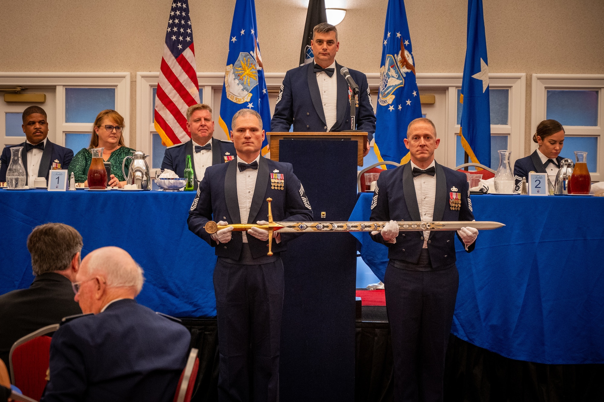 Chief Master Sgt. Gregg Gow, serving as Chief Master Sergeant of the Mess, addresses the audience from the podium during the OSI Order of the Sword ceremony, July 31, 2024, at Marine Corps Base Quantico, Virginia. From right: Chief Master Sgts Charles Pipkins and James Hoy, serving as Keeper of the Sword and Guardian of the Sword respectively, carefully place the hand-painted OSI sword in its place.