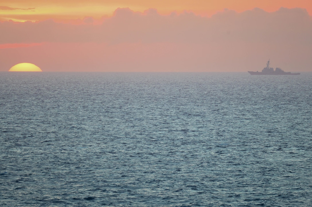 A military ship steams across the ocean with the sun partially hidden on the horizon creating an orange haze.