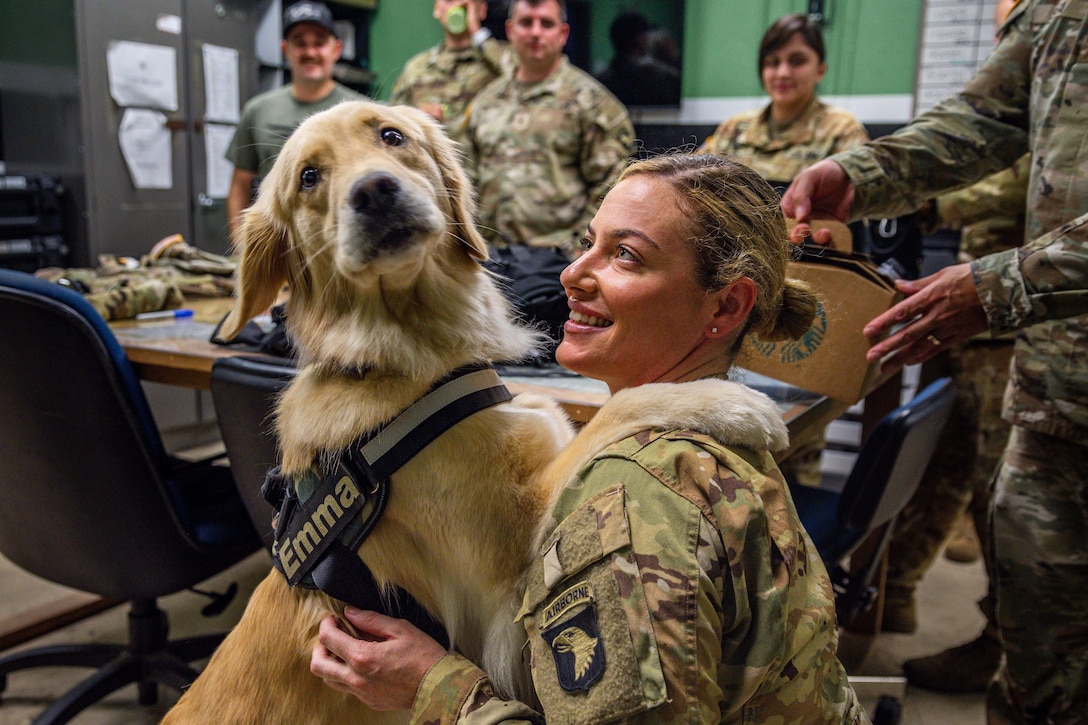 A dog with its paws on the shoulder of a smiling soldier looks at the camera while others look on in the background.