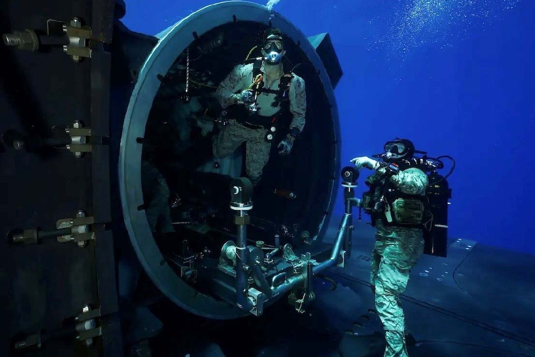 Two Marines wearing scuba gear swim underwater near a circular cement pipe close to a submarine.