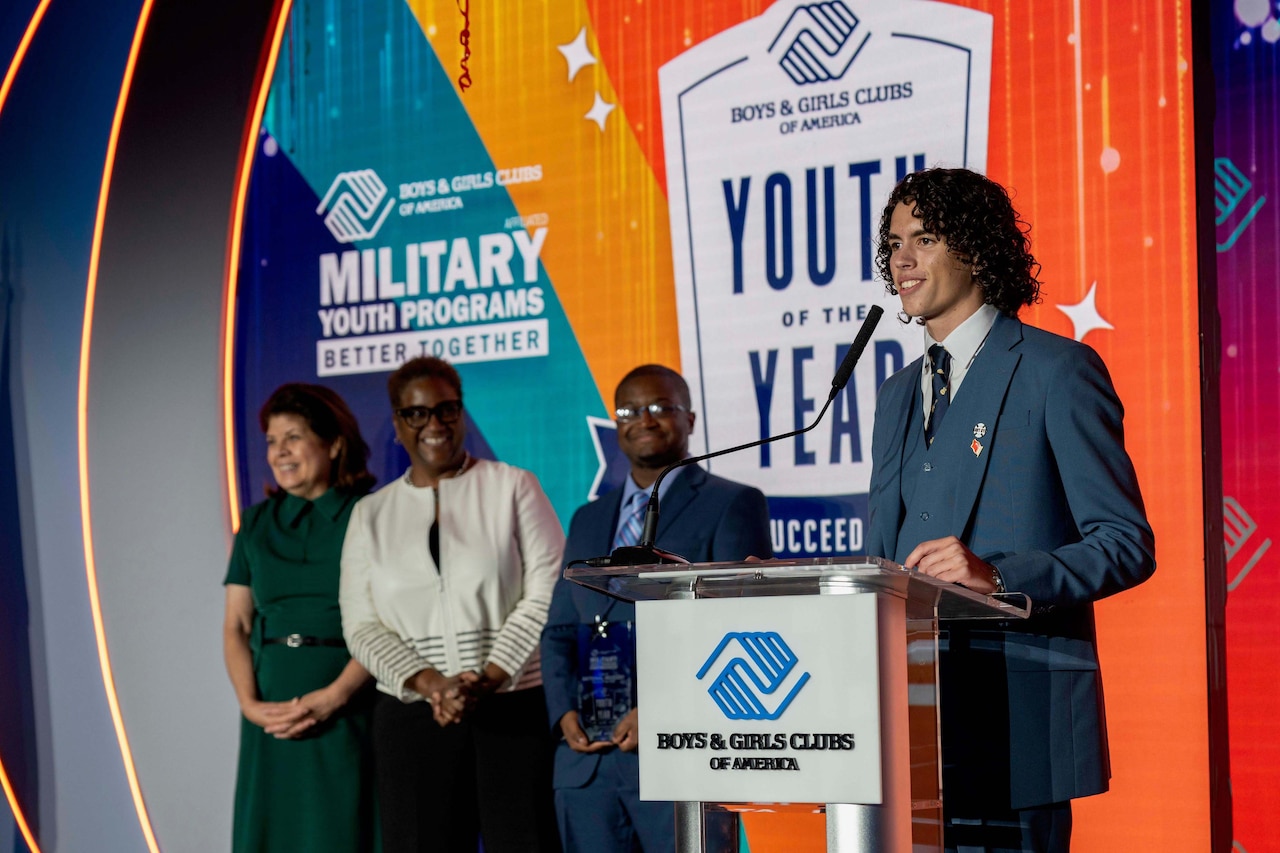 A teenage boy smiles while standing behind a lectern and three people in the background look on.