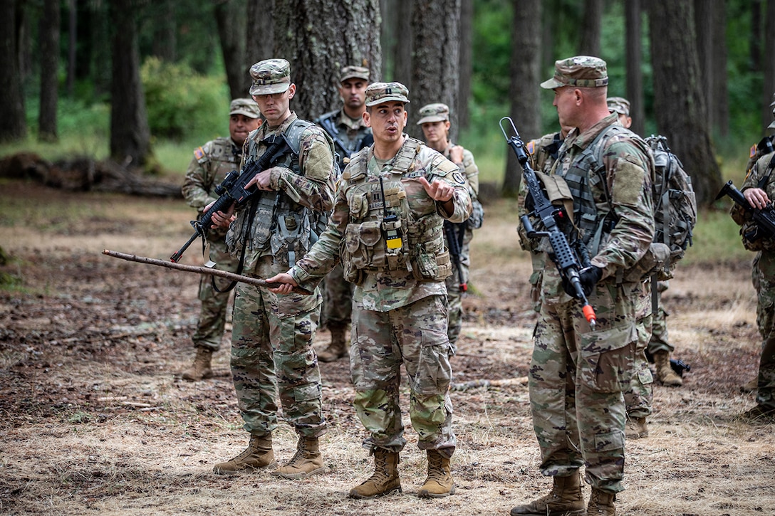 A soldier speaks to a small group of fellow soldiers holding weapons in a wooded area during daylight.