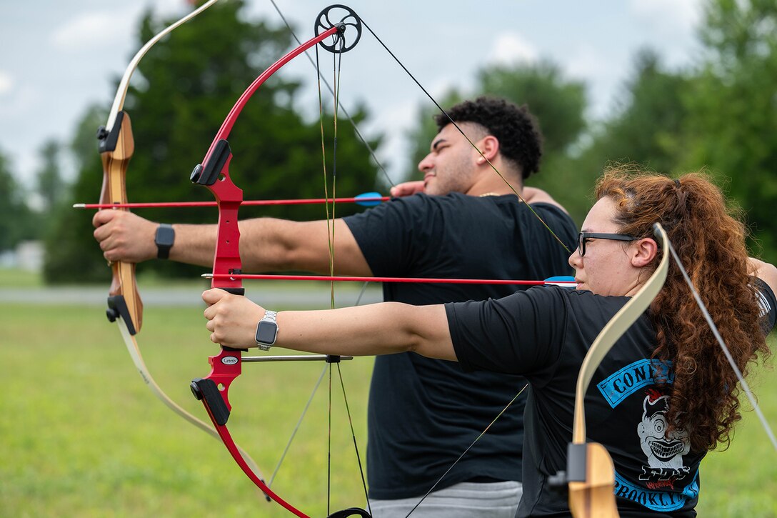 Two service members in civilian clothing practice archery during daylight.