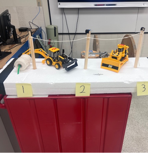 Heavy equipment models are on a table with a mock up of telephone poles and wires.