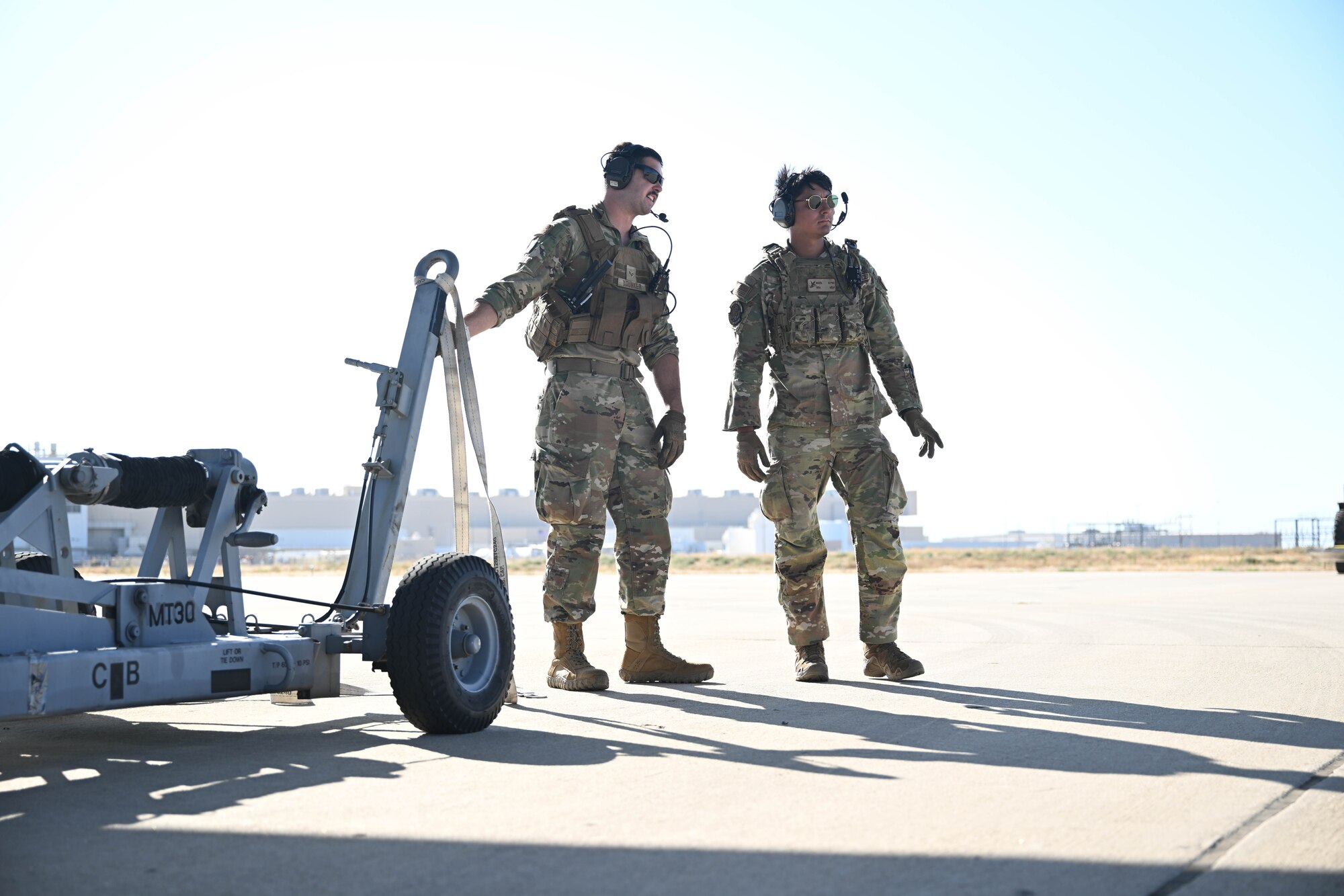 Two men in uniform stand with equipment on a flightline during the day.