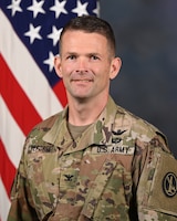 An Army colonel poses for an official portrait photo wearing an Army combat uniform (camouflage) with a U.S. flag in the background.