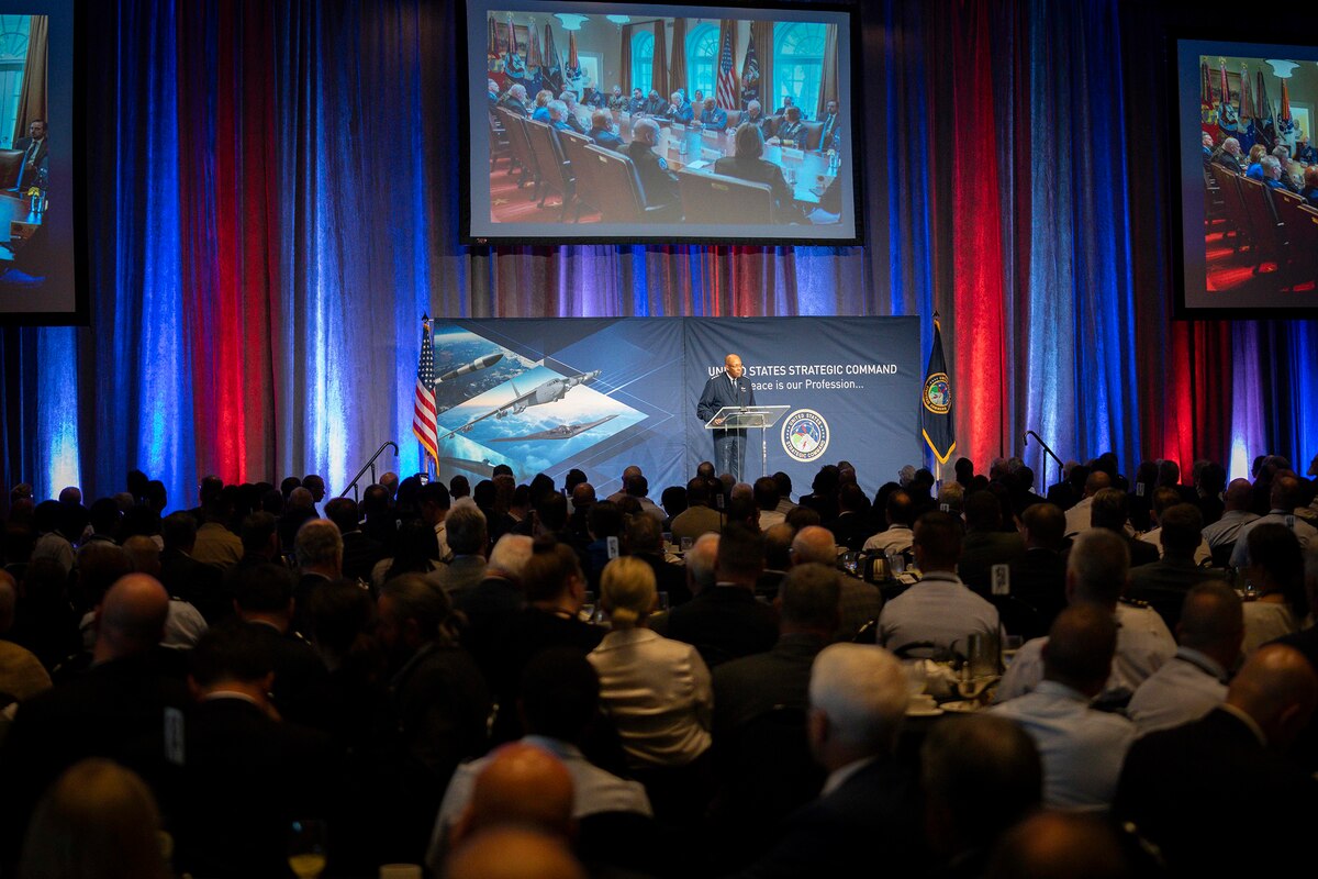 A seated audience, shown from behind, faces a general speaking into a microphone on a stage with large screens hanging overhead.